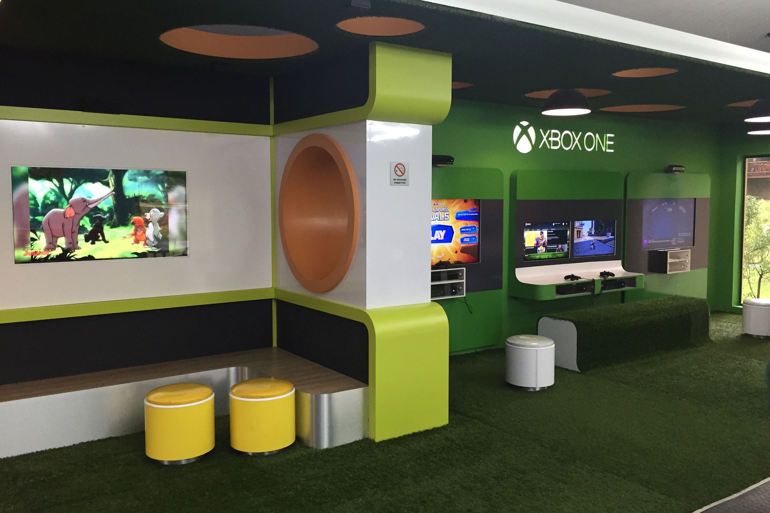 A gaming area with Xbox One consoles, a large screen, and a green and white color scheme.
