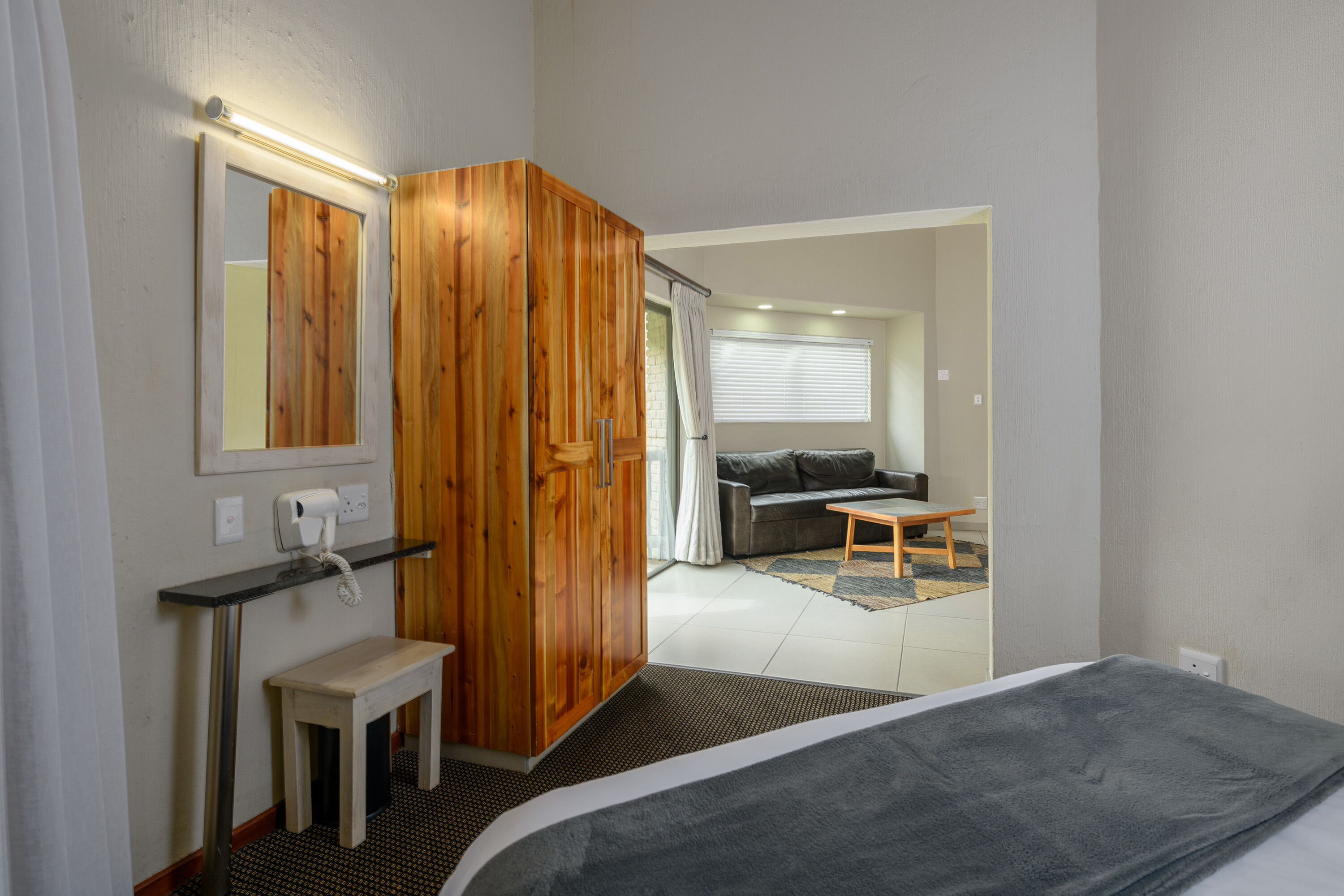 A hotel room with a bed, a wooden wardrobe, a small table, a mirror, and a bathroom with a shower curtain.