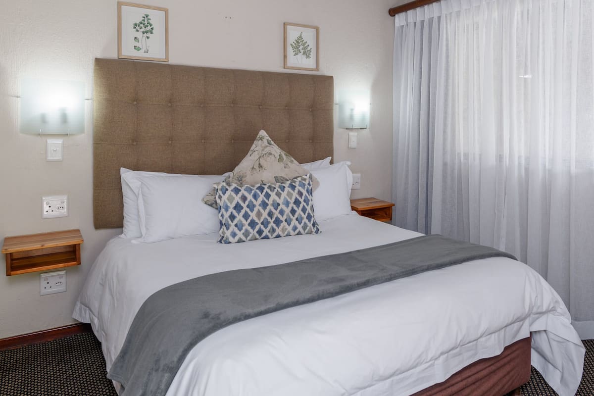 A neatly made bed with a grey blanket, white pillows, and a decorative pillow. There are two bedside tables with lamps, a wooden nightstand, and a wall-mounted light fixture.