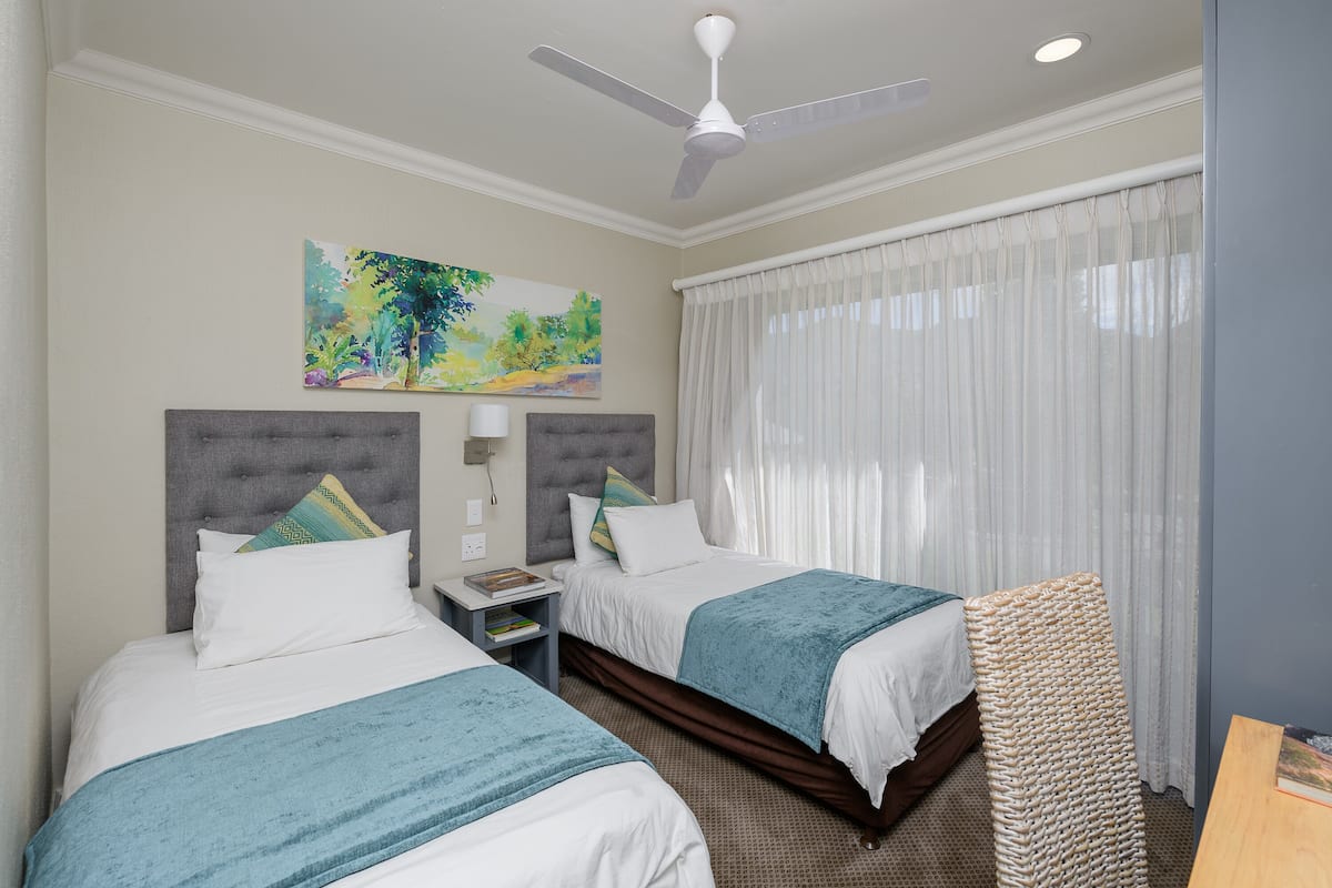 A hotel room with two beds, a ceiling fan, a painting on the wall, and a window with curtains.
