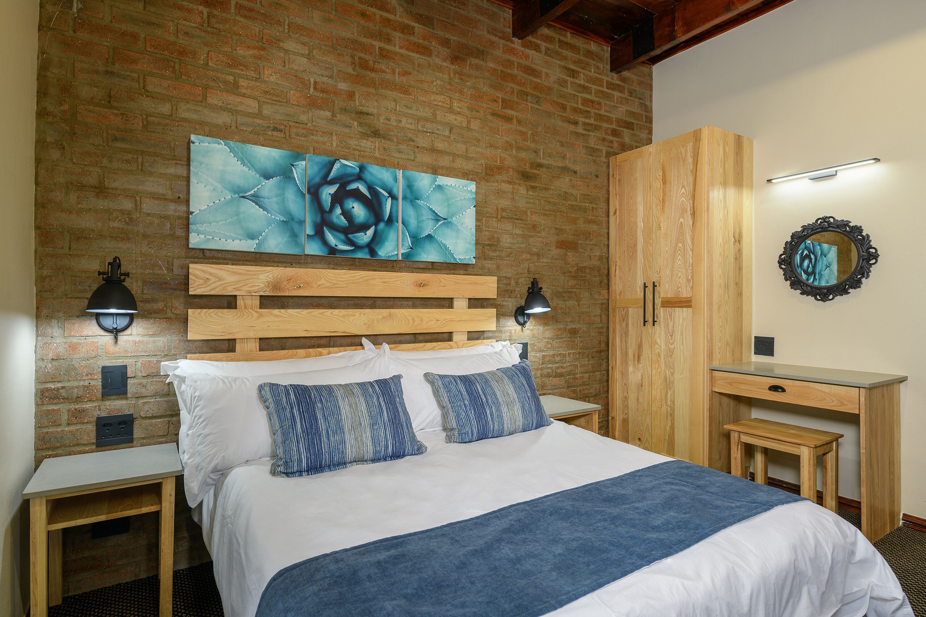 Habitación de hotel con pared de ladrillo, cama con ropa de cama blanca y almohadas azules, cabecero de madera, mesita de noche con lámpara, escritorio de madera con espejo y una obra de arte azul en la pared.