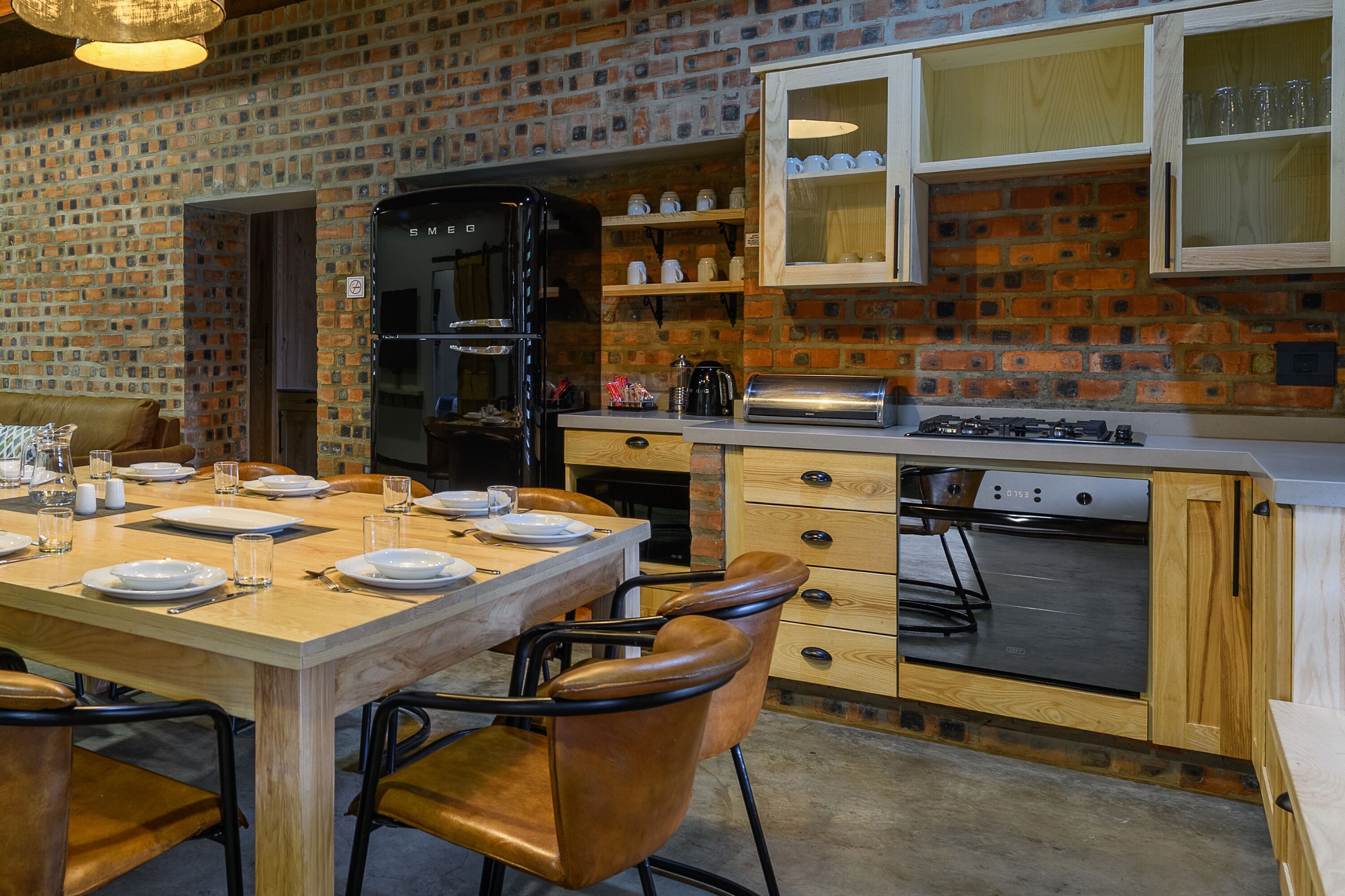 A kitchen with a brick wall, wooden cabinets, a black refrigerator, a stove, and a dining table set with plates and glasses.