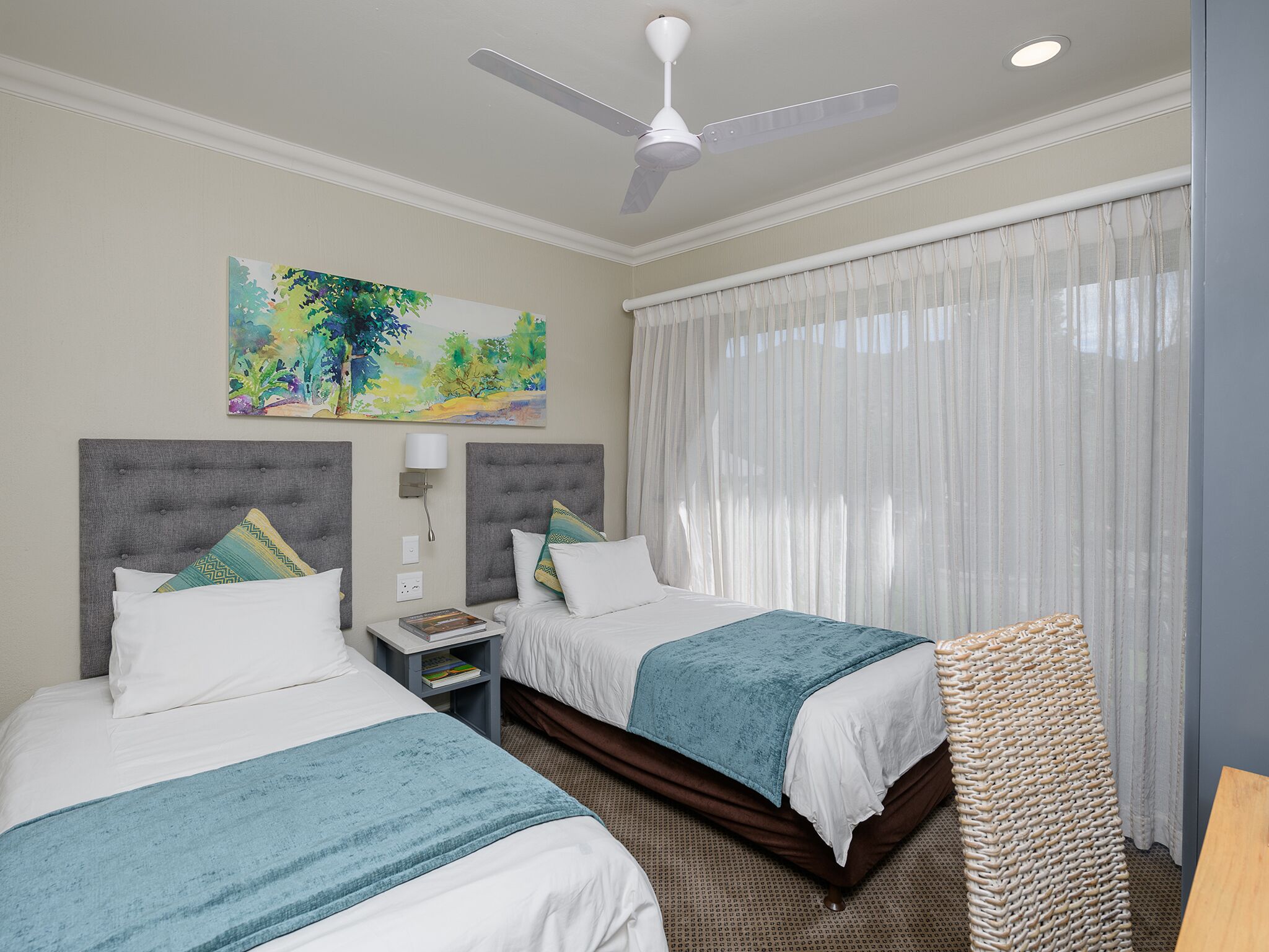 Quarto de hotel com duas camas, um ventilador de teto, um quadro, uma mesa de cabeceira e uma janela grande com cortinas.