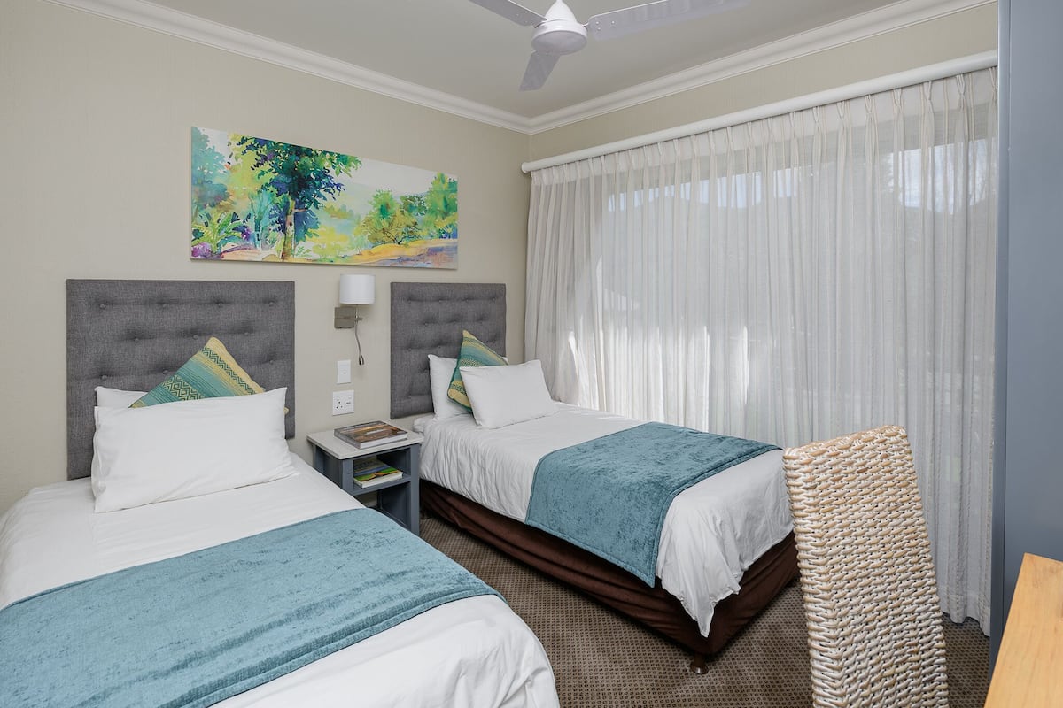 A hotel room with two beds, a ceiling fan, a painting, a nightstand, and a large window with curtains.