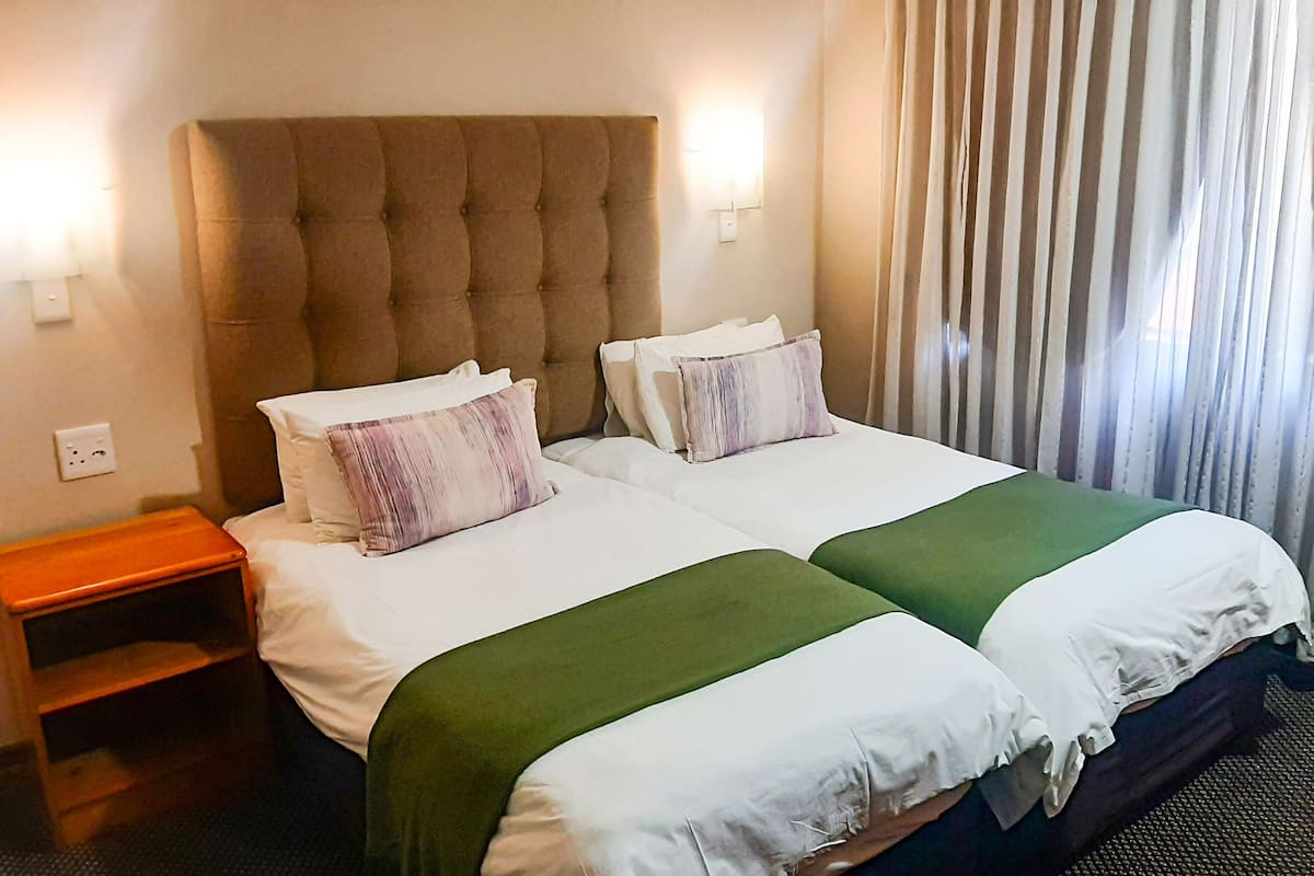 A hotel room with two beds, a wooden nightstand, and a window with curtains.