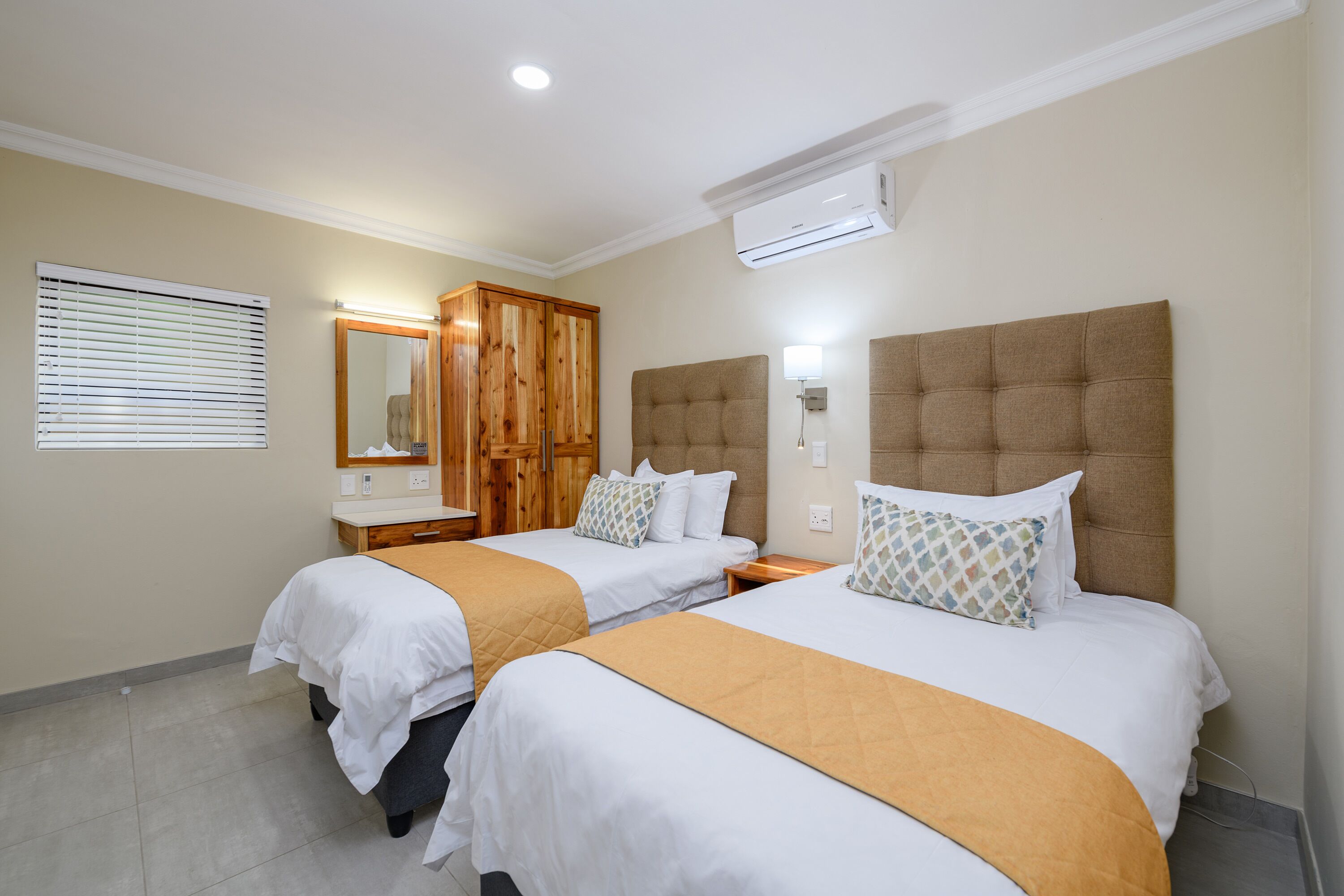 A hotel room with two beds, a wooden wardrobe, a mirror, and an air conditioning unit.