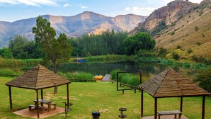 Un camping tranquilo con dos quioscos de madera, mesas de picnic y un pequeño muelle junto a un lago apacible, rodeado de exuberante vegetación y montañas al fondo.