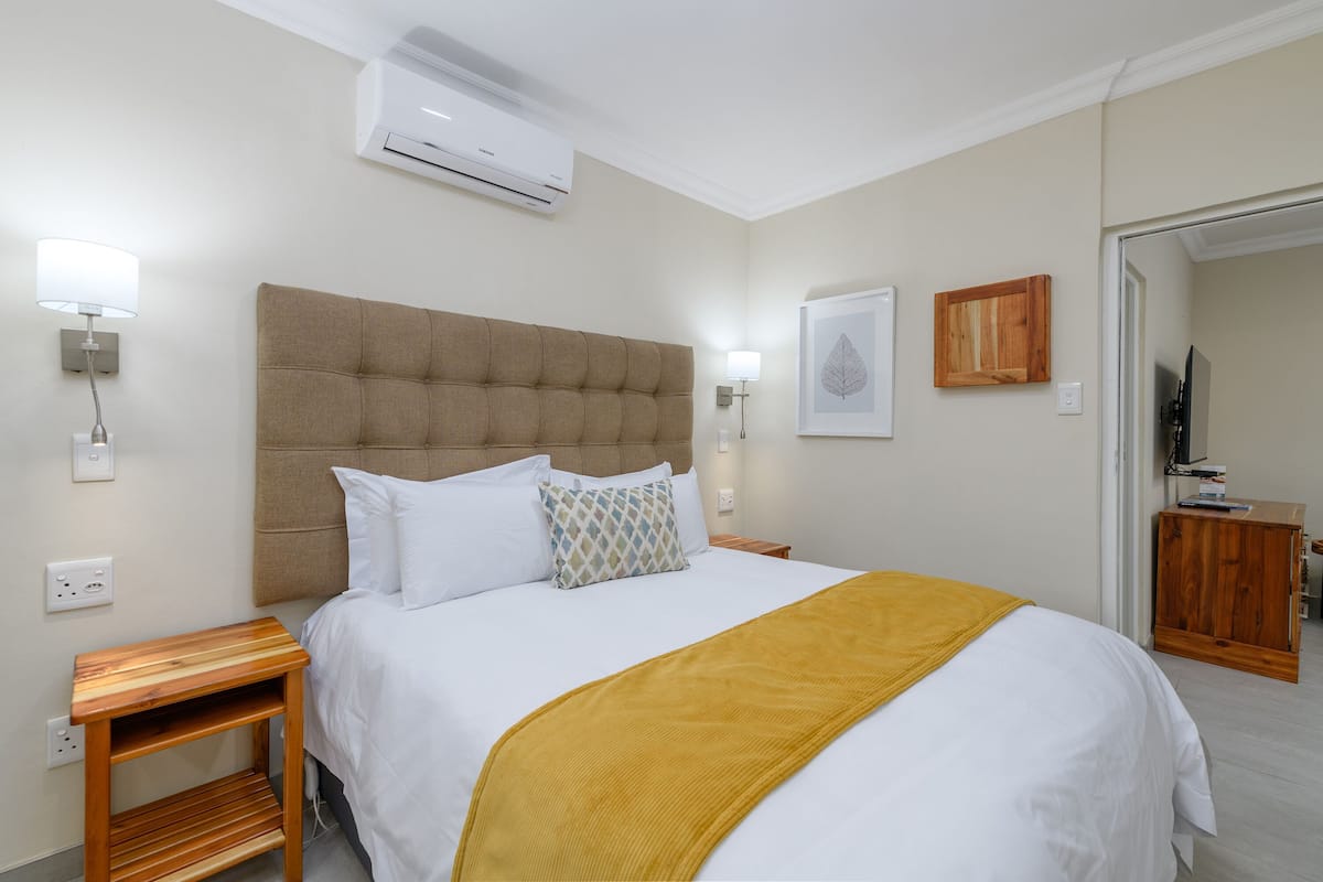 A neatly made bed with a headboard, a bedside table, a wall-mounted air conditioner, a framed picture, and a wooden cabinet.