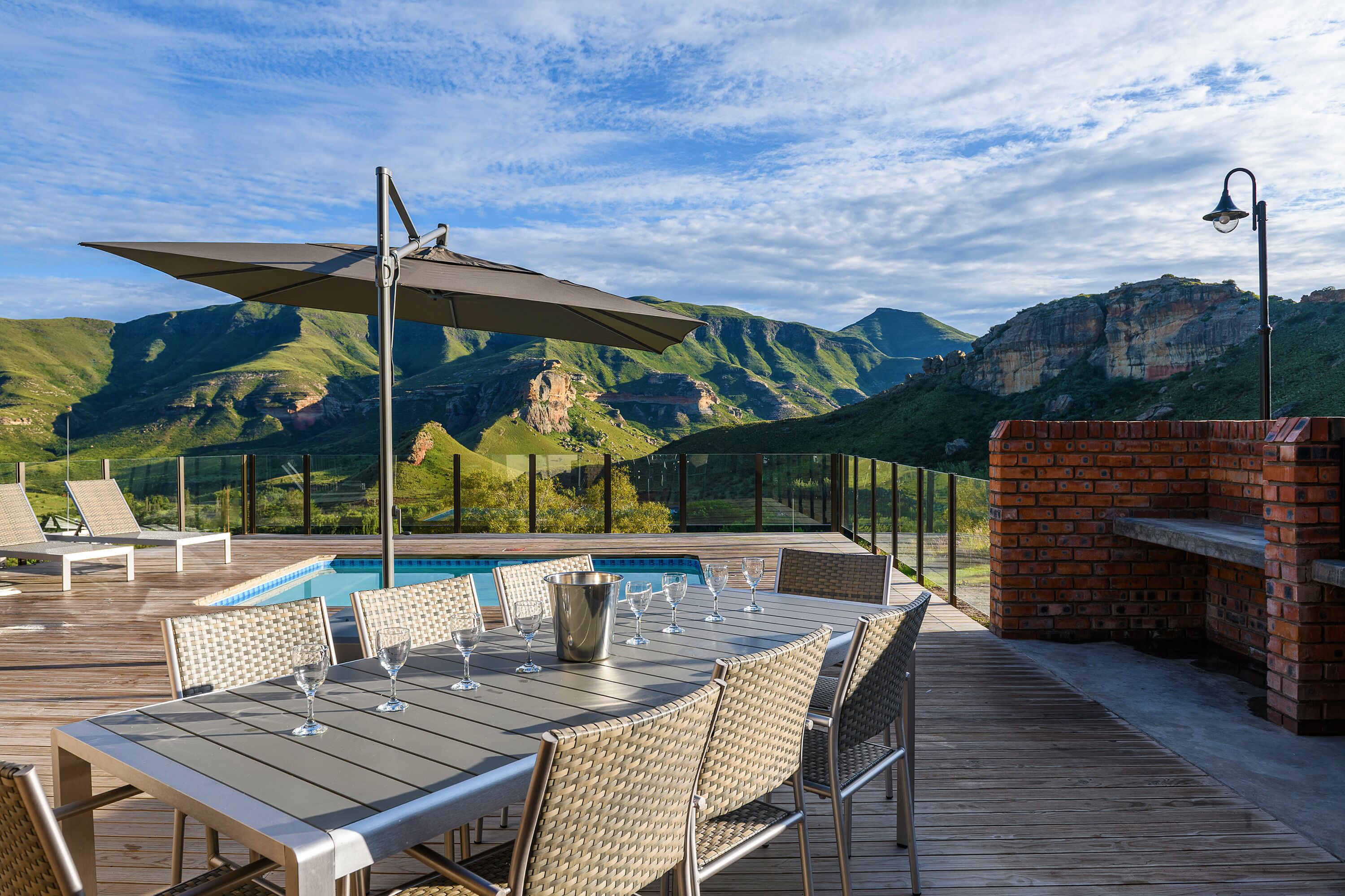 A deck with a pool, outdoor seating, and a brick barbecue area, overlooking a mountainous landscape.