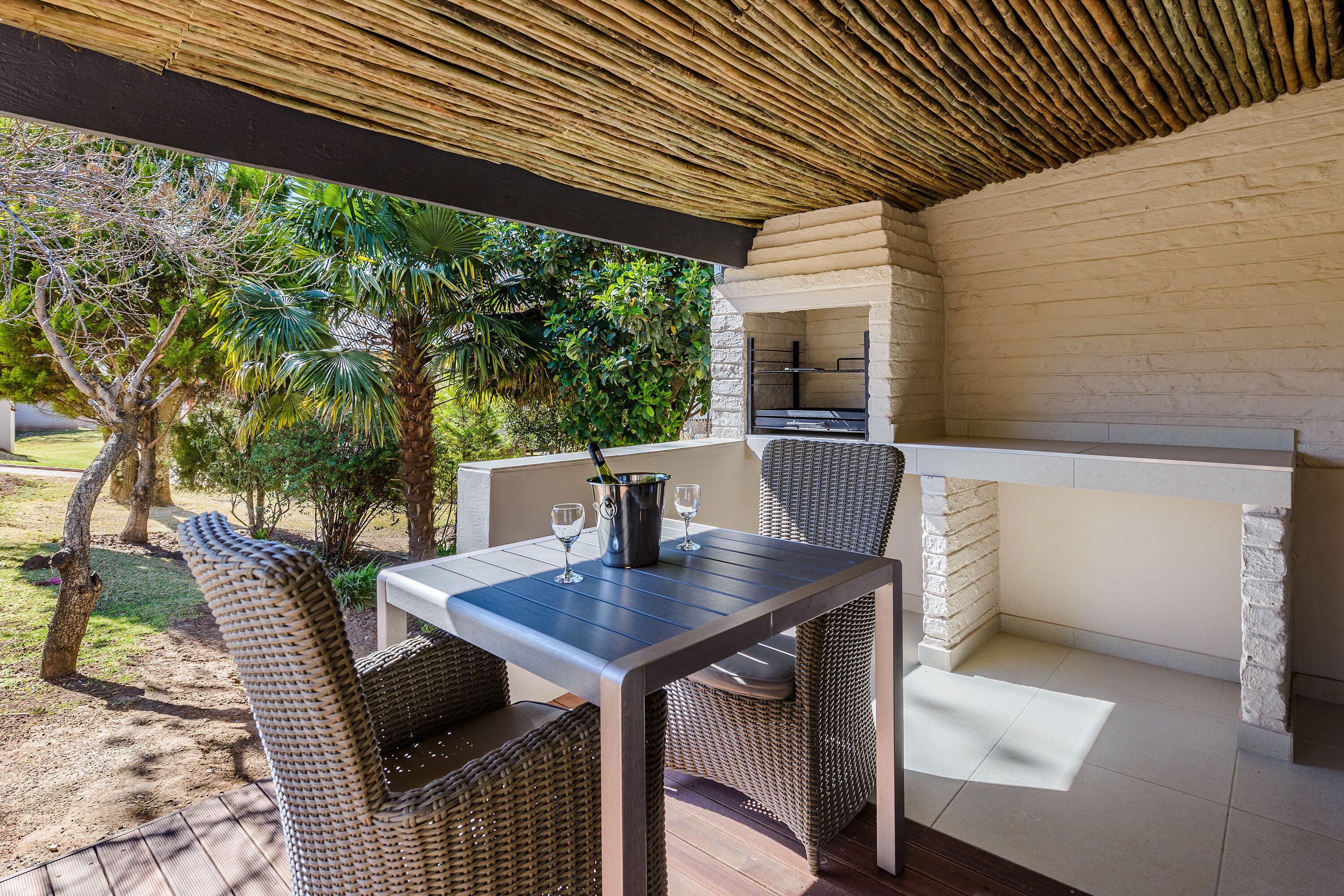 An outdoor patio with wicker furniture, a table with a bottle and glasses, and a built-in grill.