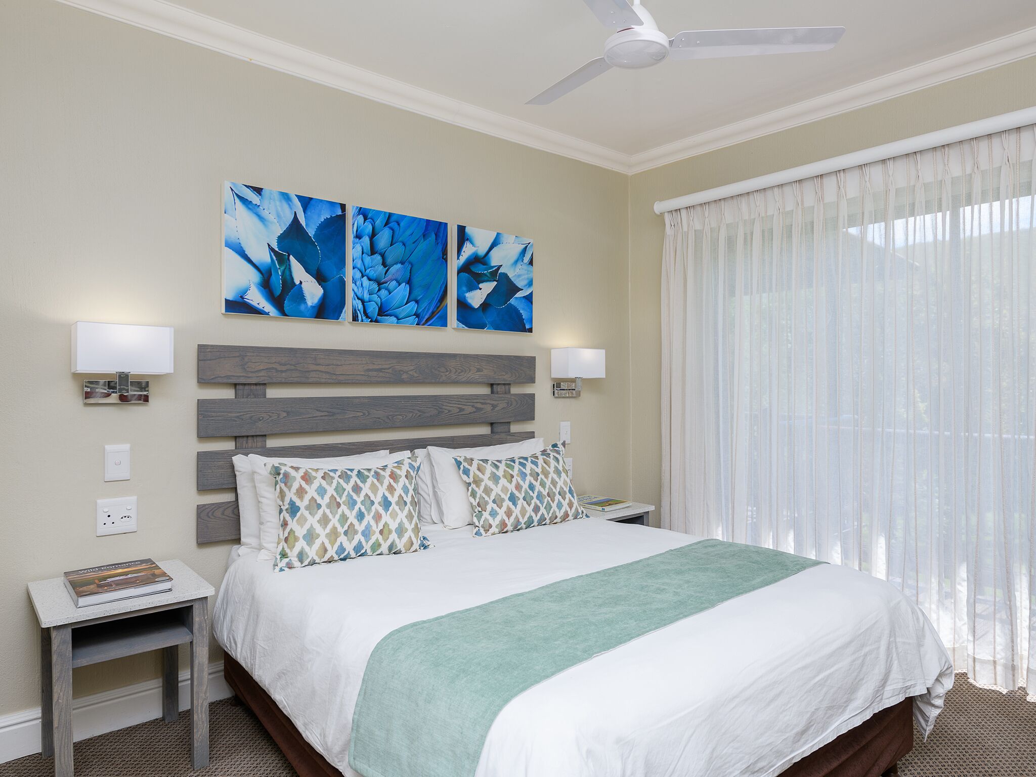 Quarto de hotel com uma cama, mesa de cabeceira, ventoinha no teto e obras de arte na parede.