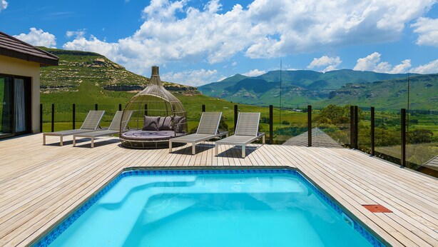 Una terraza de madera con piscina, sillas de descanso y jacuzzi, con vistas a un paisaje montañoso.