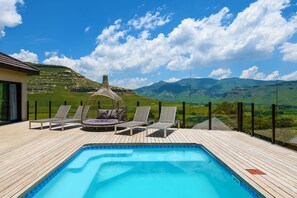 Una terraza de madera con piscina, sillas de descanso y jacuzzi, con vistas a un paisaje montañoso.