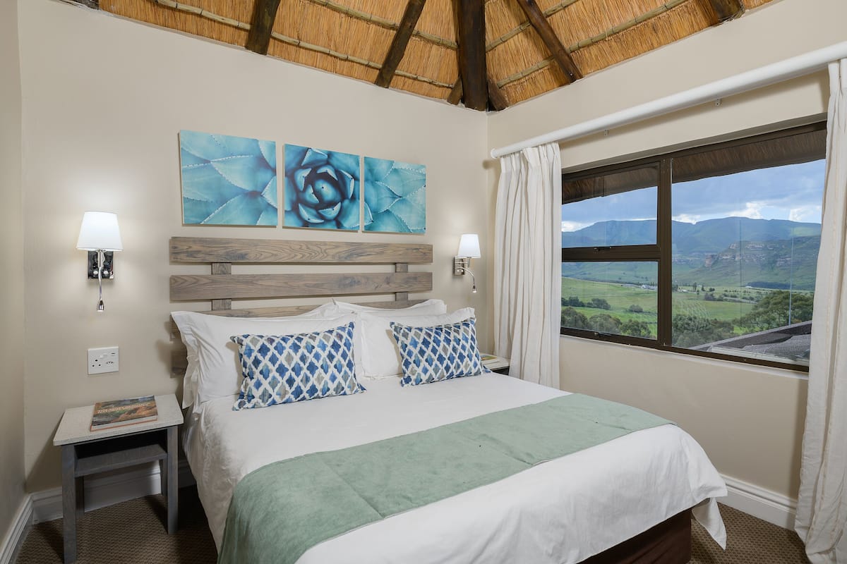 A bedroom with a bed, two wall-mounted lamps, a nightstand, a window with a view of the countryside, and a wooden headboard.