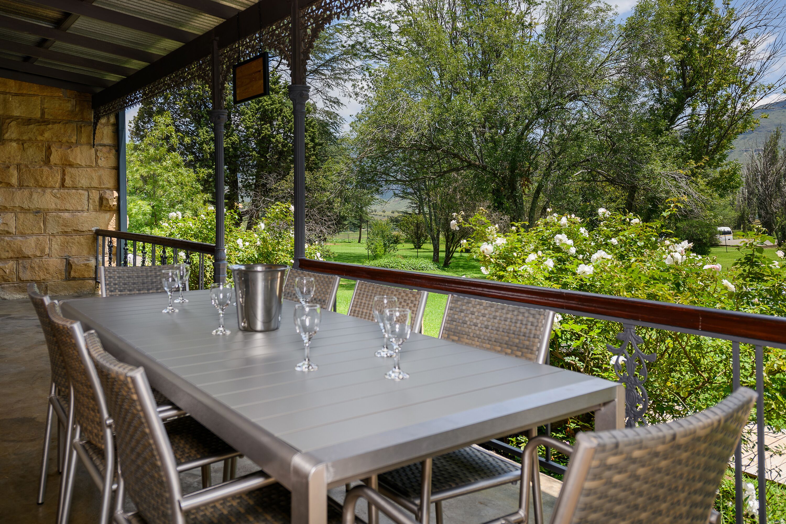 Une terrasse extérieure avec une table dressée pour le repas, entourée de chaises en osier, donnant sur un jardin luxuriant et sur un paysage montagneux en toile de fond.