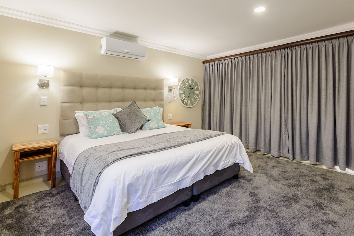 A bedroom with a large bed, a wall clock, an air conditioner, and a window with curtains.