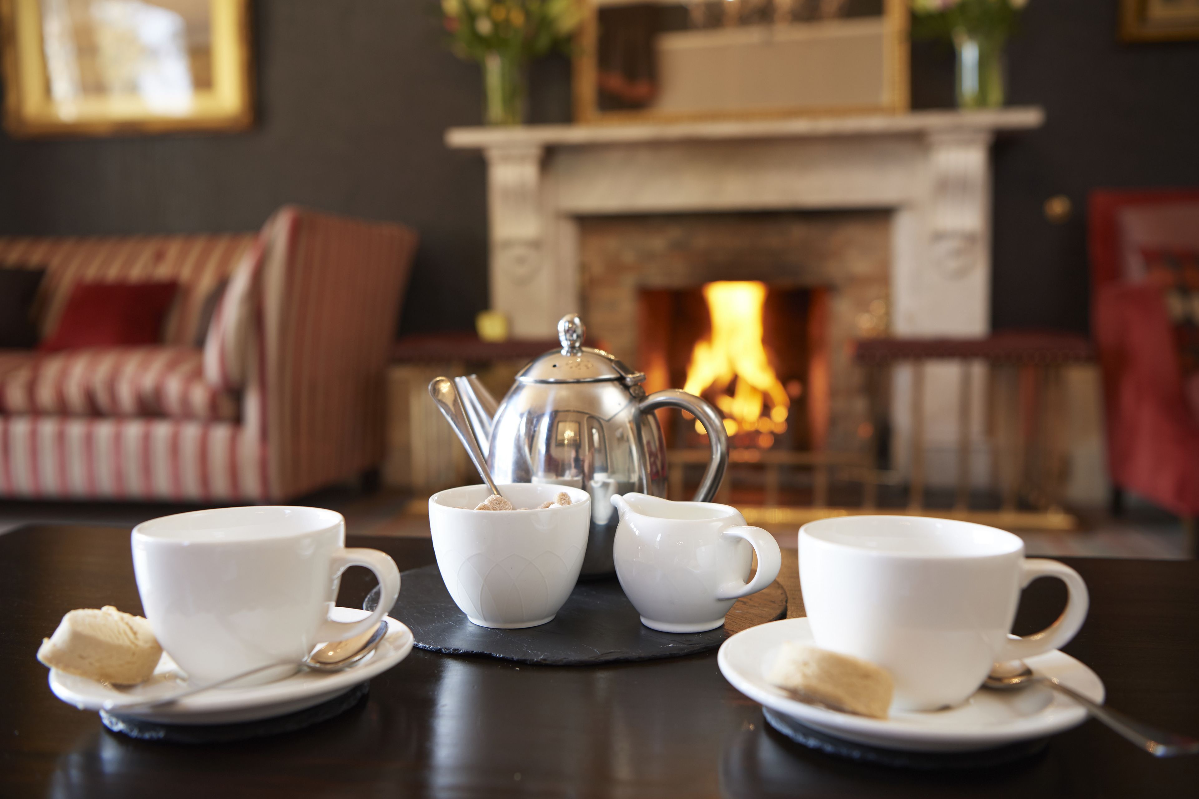 A table set with white cups and saucers, a silver teapot, and a sugar bowl, with a fireplace in the background.