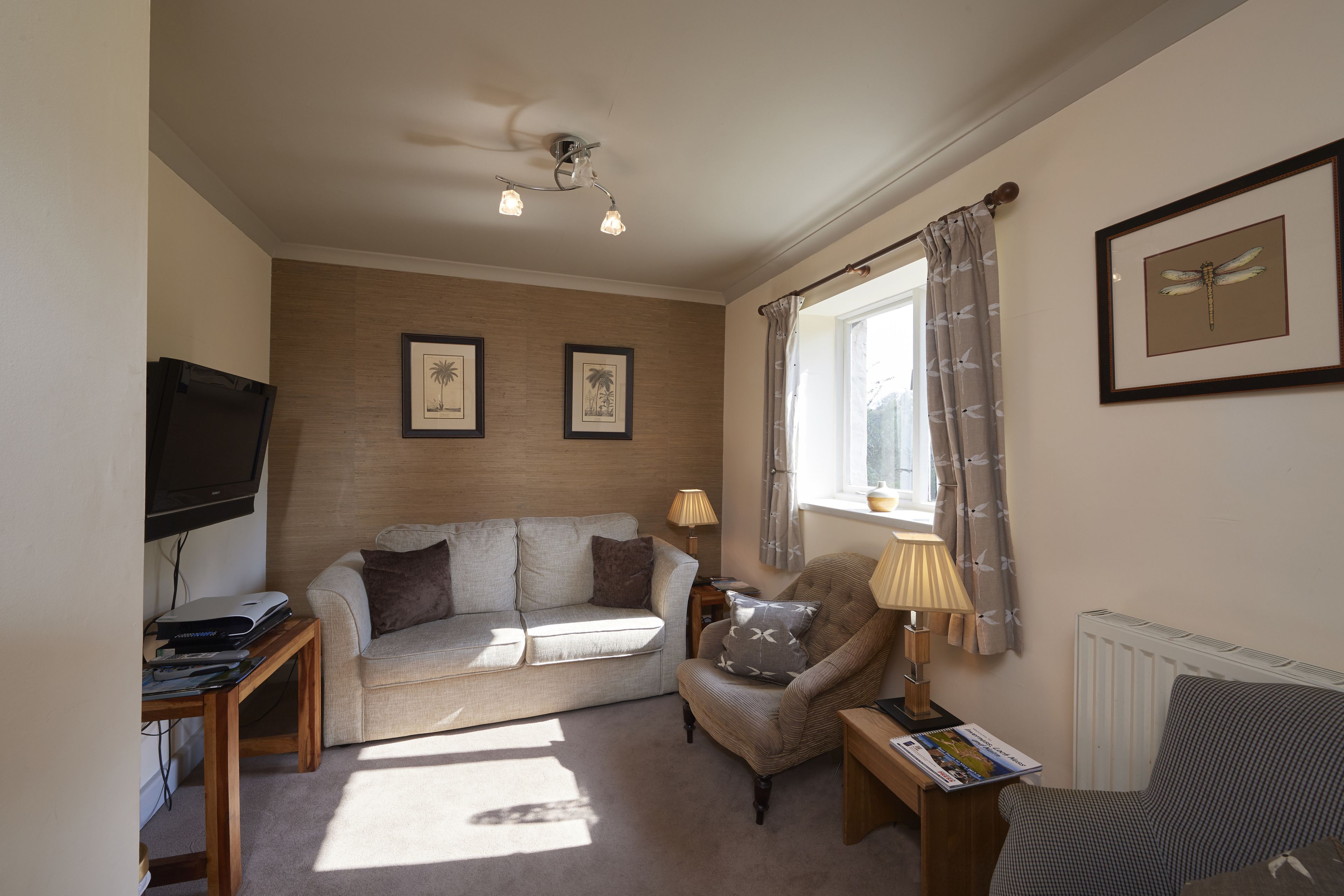 A living room with a sofa, armchair, and a TV on a stand. There are framed pictures on the wall and a window with curtains.