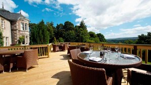 A wooden deck with wicker furniture and a glass table set for dining, overlooking a scenic landscape.