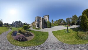 A historic stone building with a curved facade, surrounded by well-manicured gardens and a gravel driveway.