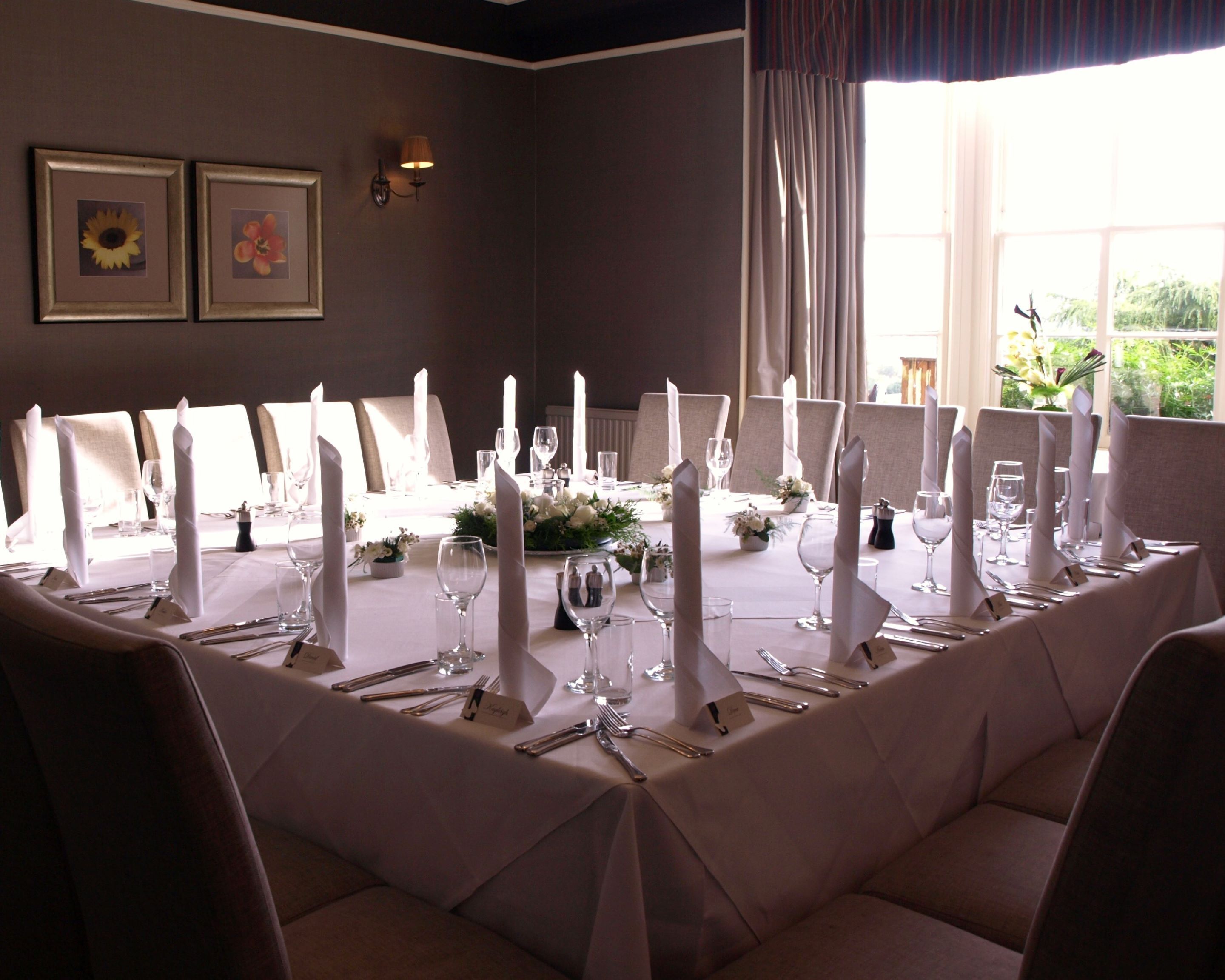 A formal dining room with a long table set for a meal, framed pictures on the wall, and a window with curtains.