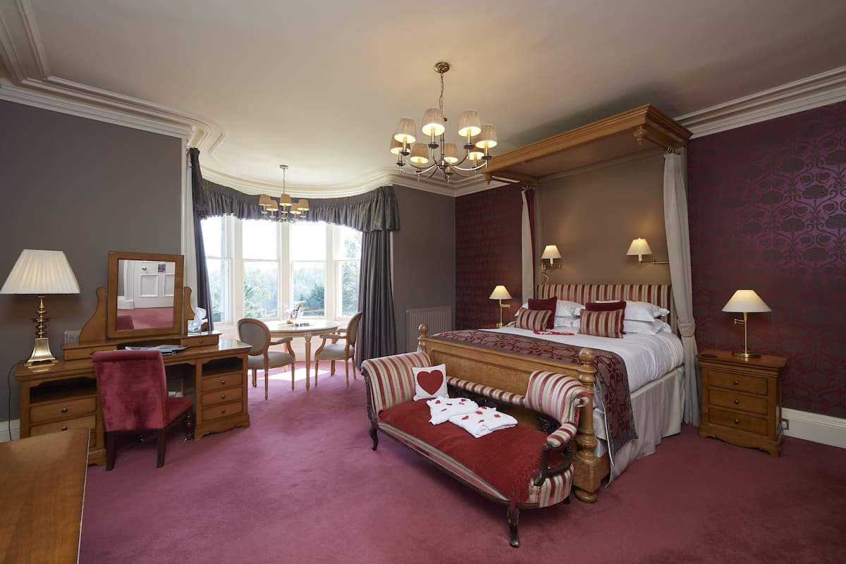 A spacious bedroom with a four-poster bed, a sitting area with a bench, a desk, and a chair.