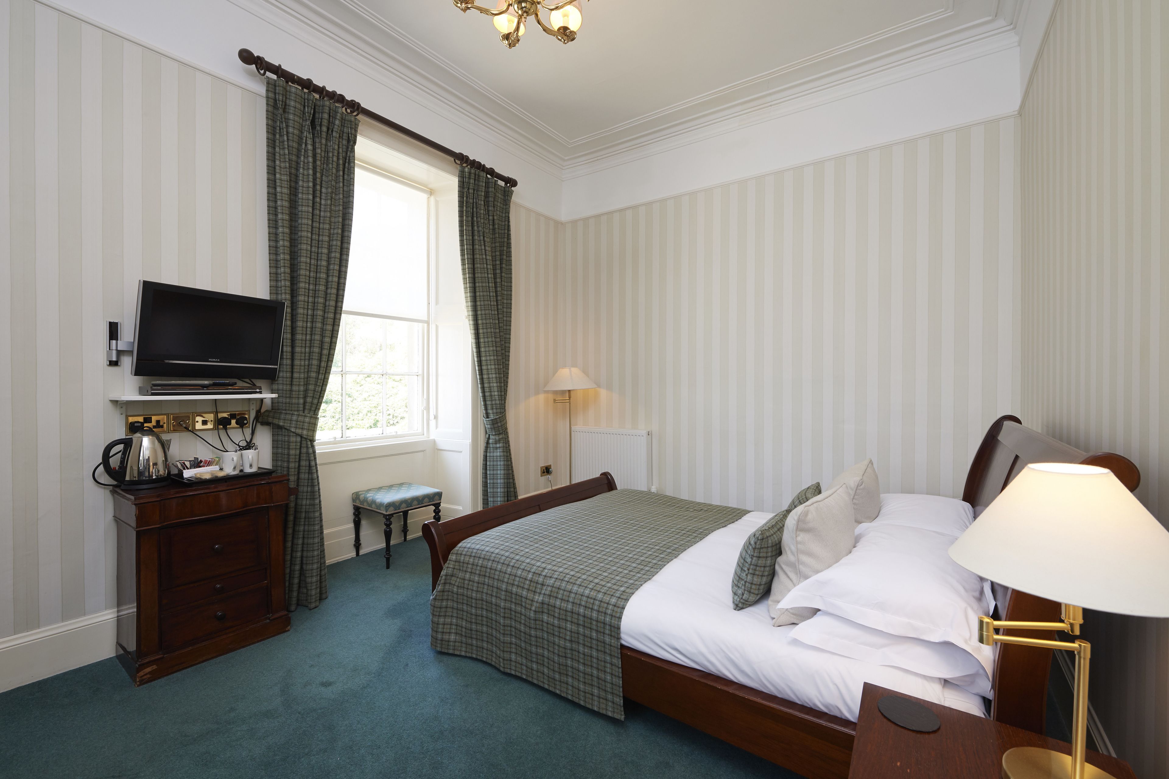 A neatly arranged bedroom with a bed, bedside table, lamp, television, dresser, and a window with curtains.