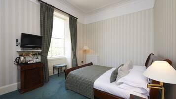 A neatly arranged bedroom with a bed, bedside table, lamp, television, dresser, and a window with curtains.