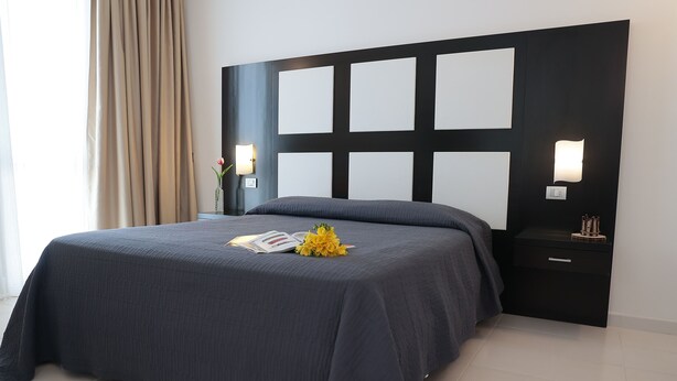A modern bedroom with a large bed, a nightstand with a lamp, and a wall-mounted light fixture. - Hotel Admeto (Castelvetrano)