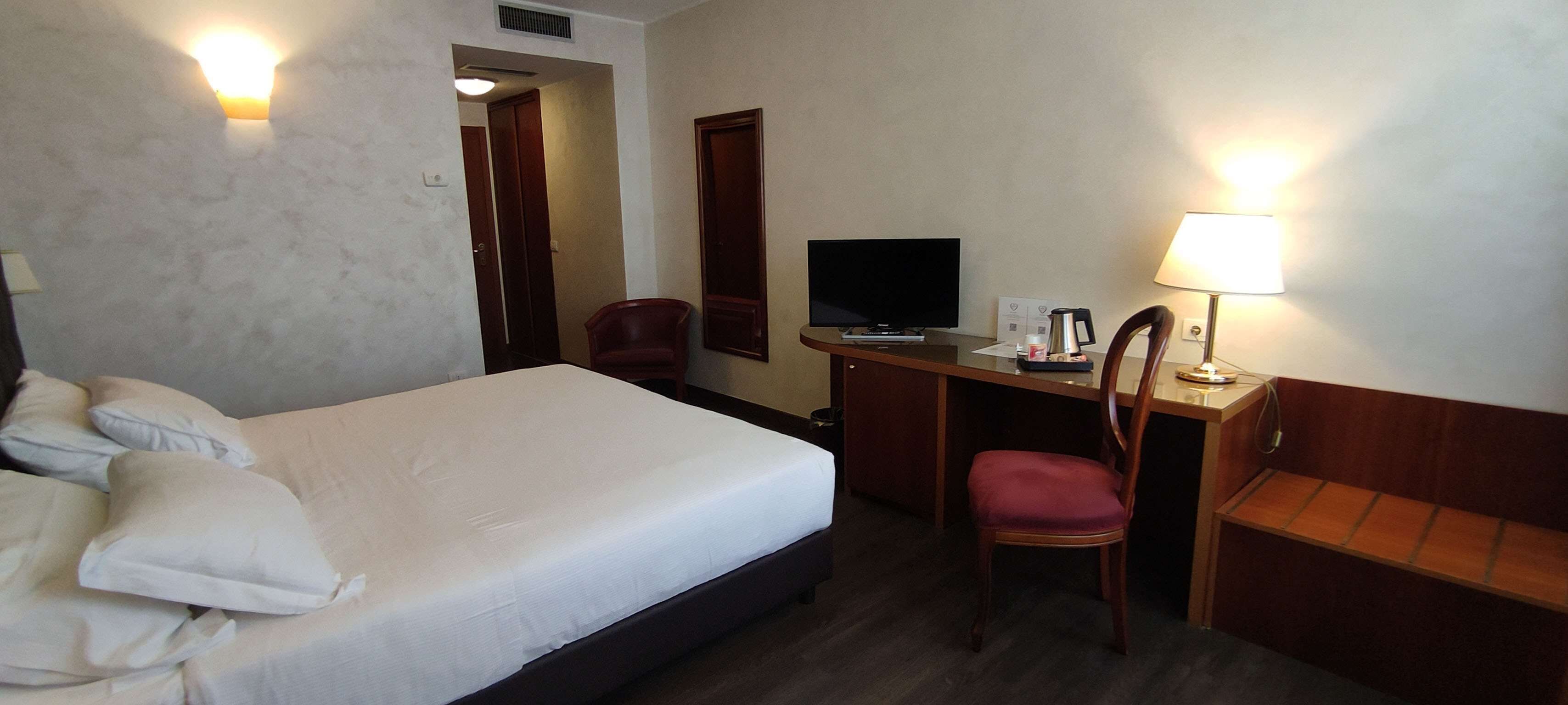 Una camera d'albergo con un letto, una scrivania con una sedia, una televisione, una lampada e un tavolino con una macchina per il caffè.