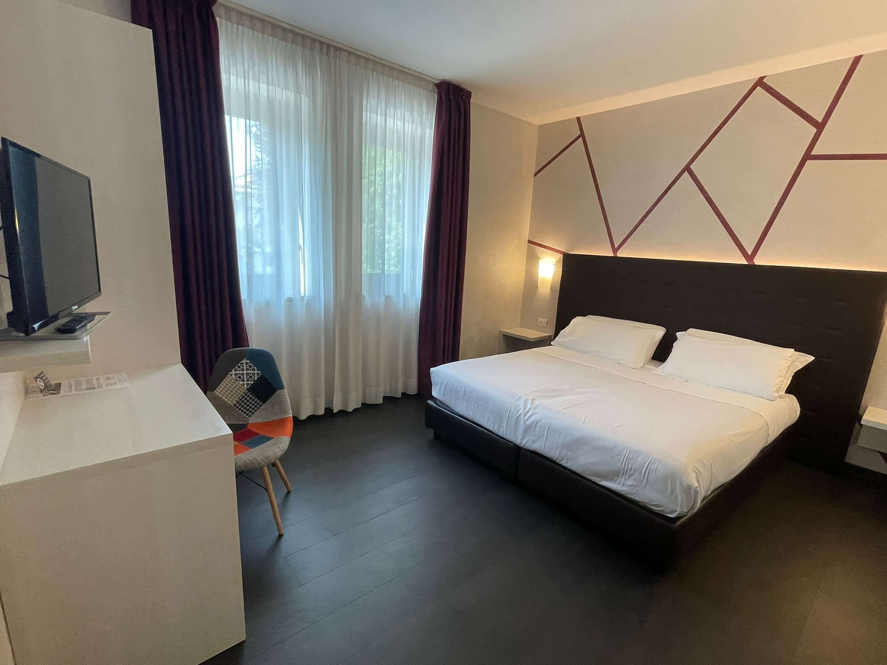A hotel room with a bed, a desk with a TV, a chair, and a window with curtains.