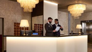 A reception area with a reception desk, two individuals conversing, a chandelier, a wall-mounted lamp, and a display of trophies. - Best Western Park Hotel Continental (San Dona di Piave)