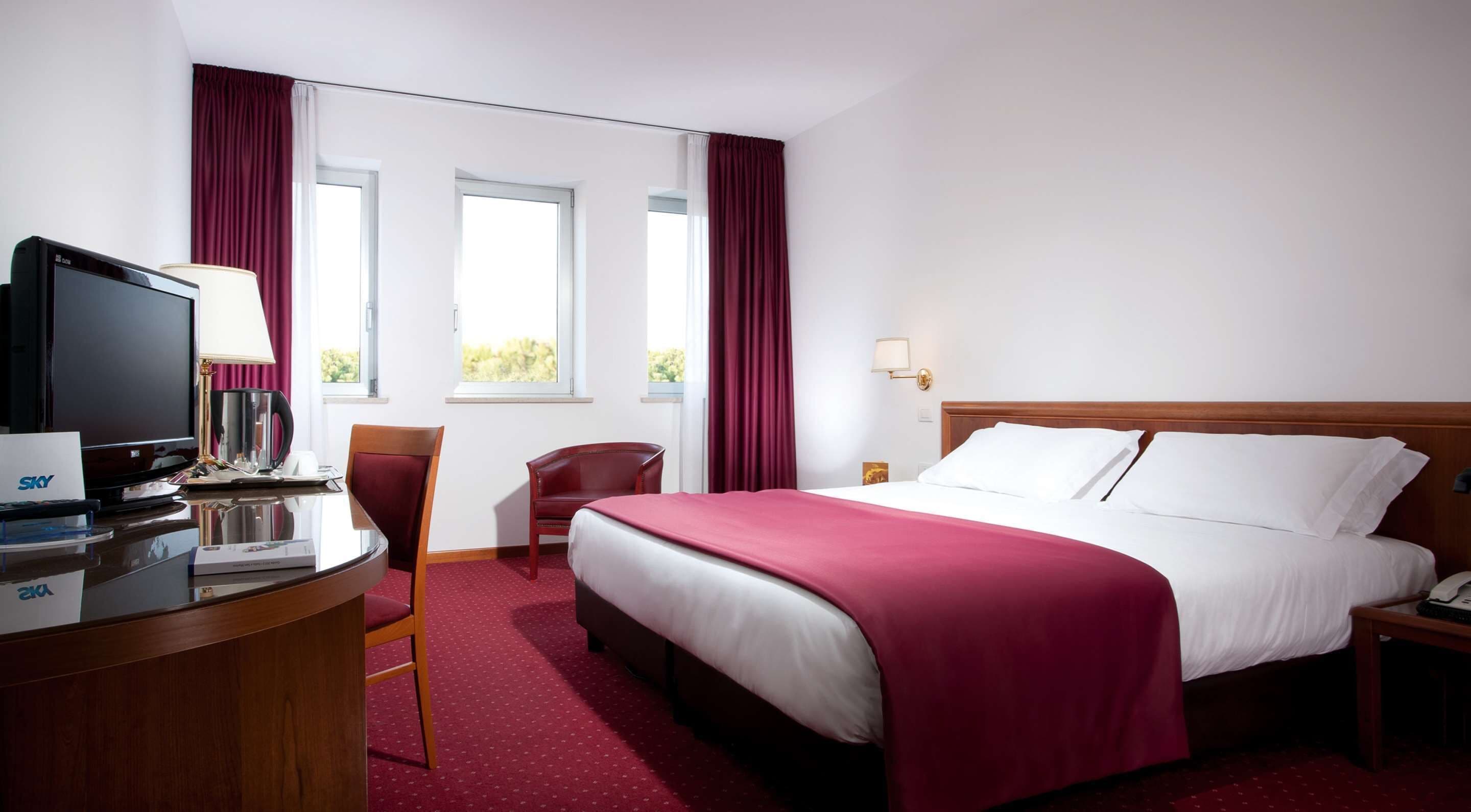 A hotel room with a bed, a desk with a TV, a chair, and a window with curtains.