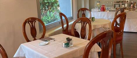 A dining area with wooden chairs and tables, a window with a view of greenery, and a buffet setup in the background.