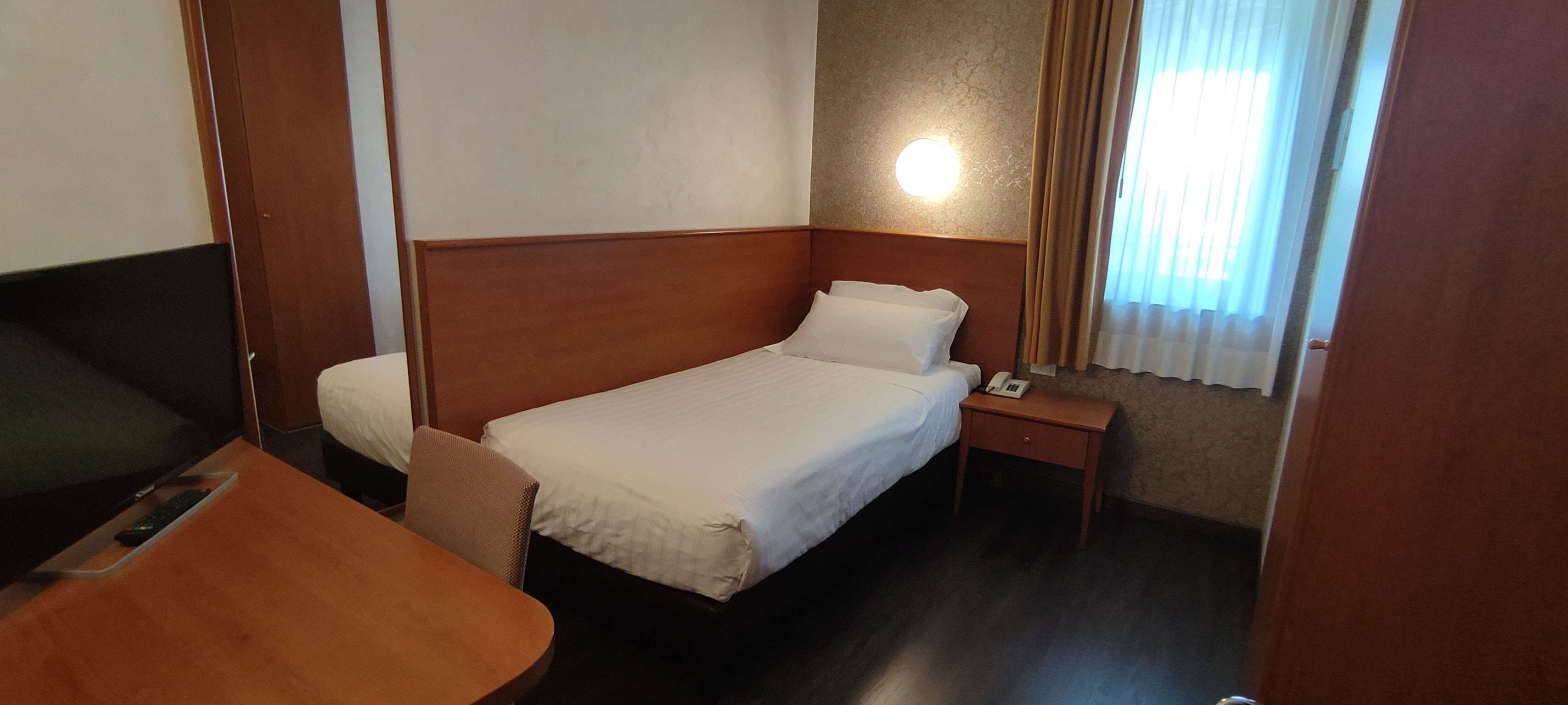Een hotelkamer met een enkel bed, een nachtkastje met lamp, een televisie en een houten bureau.