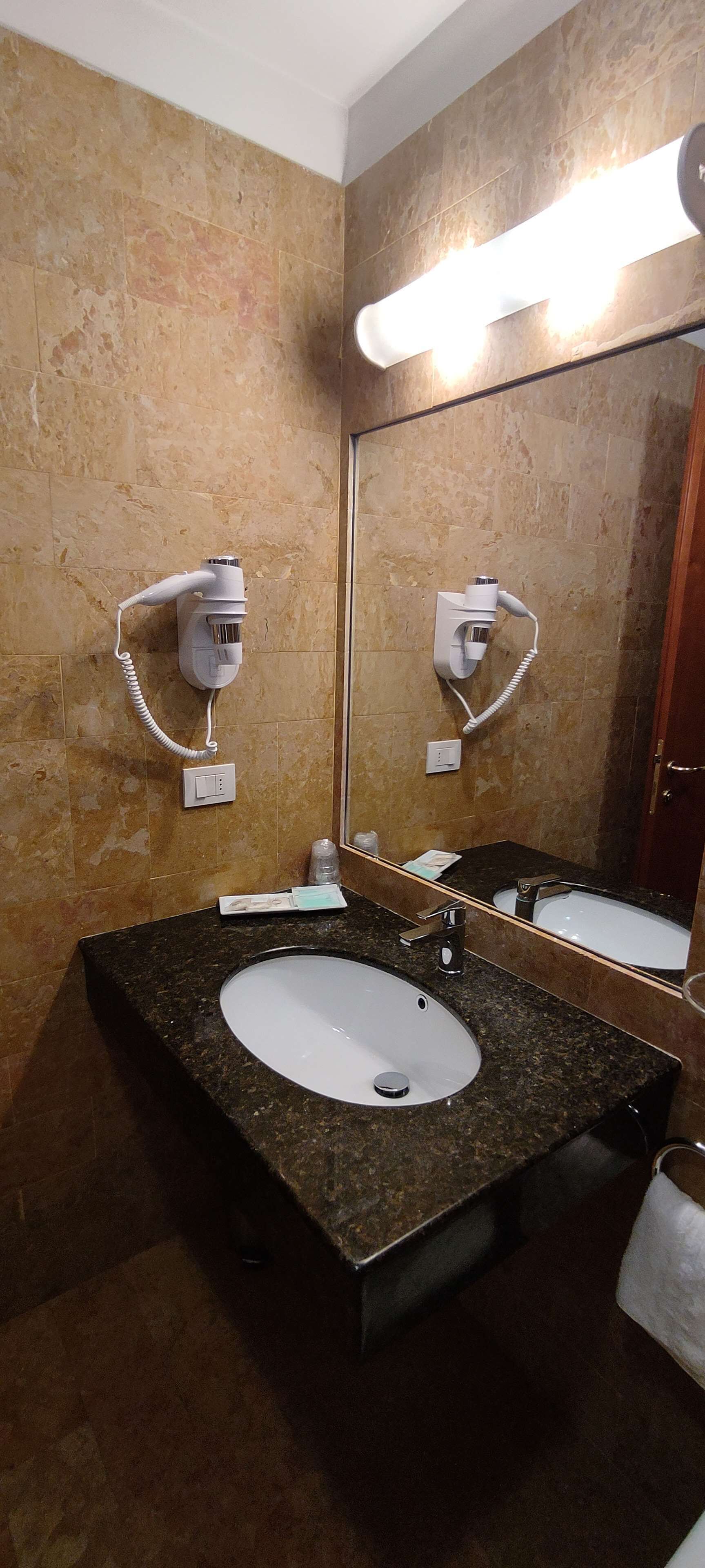 Un baño con lavamanos de encimera de granito, dos secadores de pelo montados en la pared y un espejo grande.