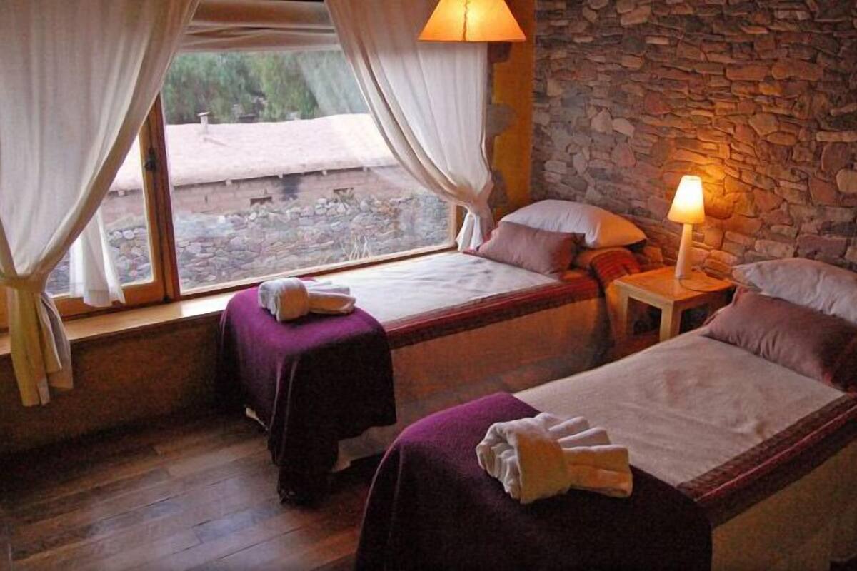 A room with two beds, a stone wall, a window with curtains, and a lamp.