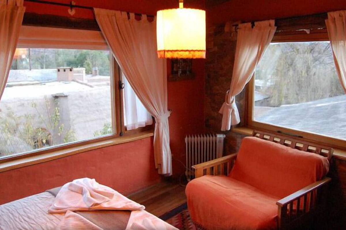 A room with a bed, a chair, a window with curtains, a radiator, and a lamp.
