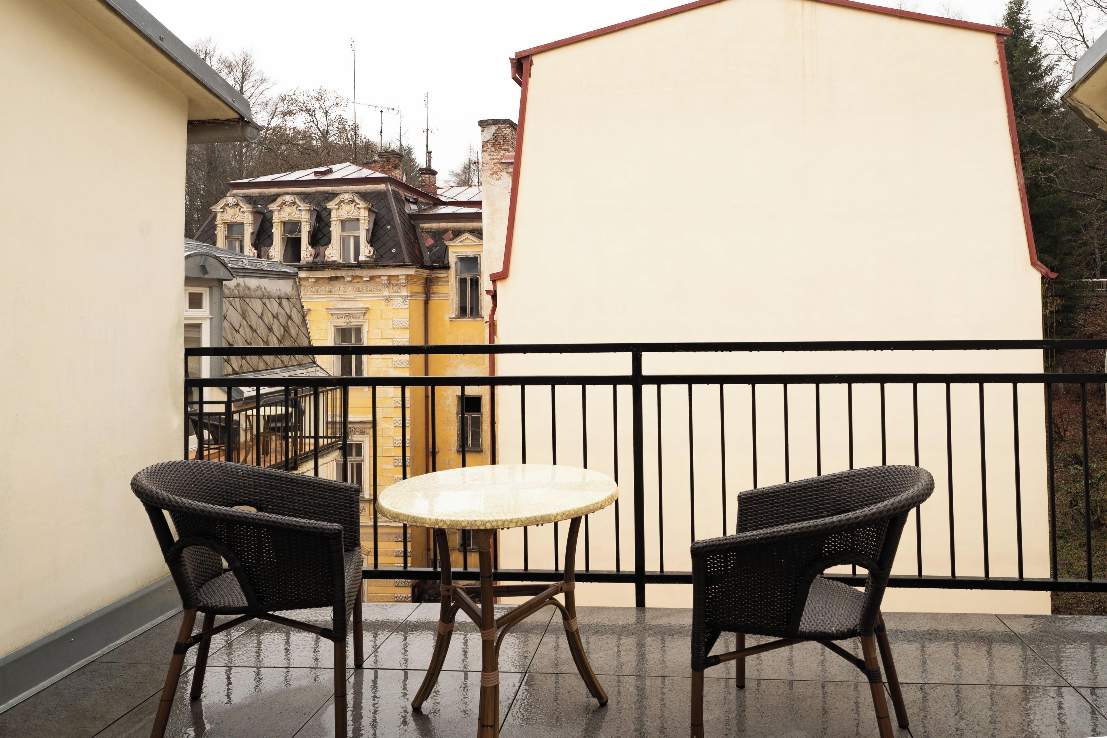 A balcony with two wicker chairs and a round table, overlooking a cityscape with historic buildings.