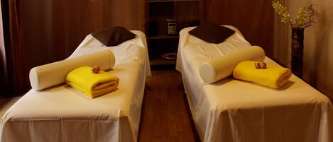 Two single beds with white linens, a wooden floor, a small shelf with items, and a vase with flowers.