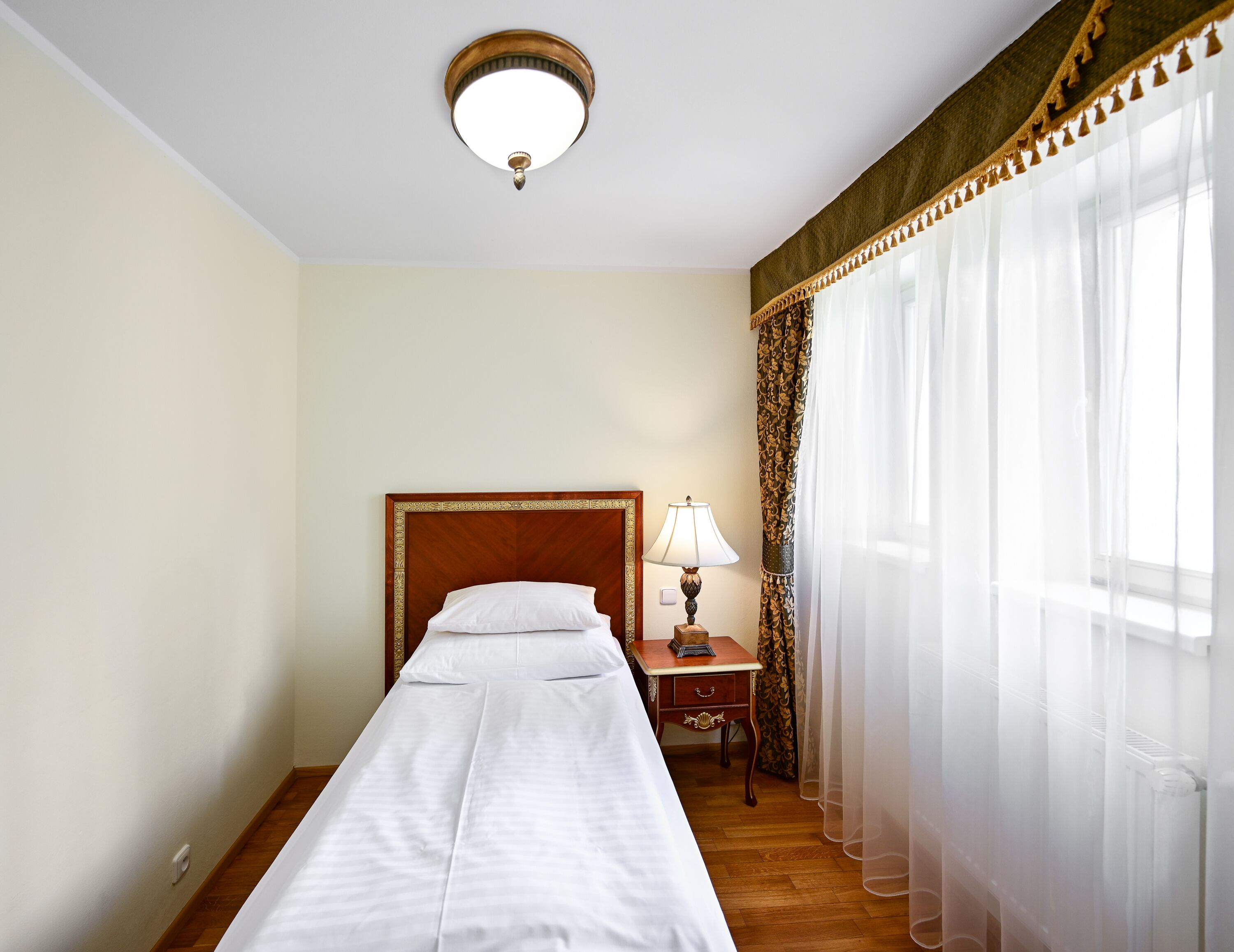 A hotel room with a single bed, a bedside table with a lamp, a window with sheer curtains, and a ceiling light fixture.