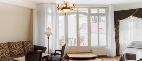 A hotel room with a sofa, a dining table, a chair, a lamp, a bed, and a large window with curtains.