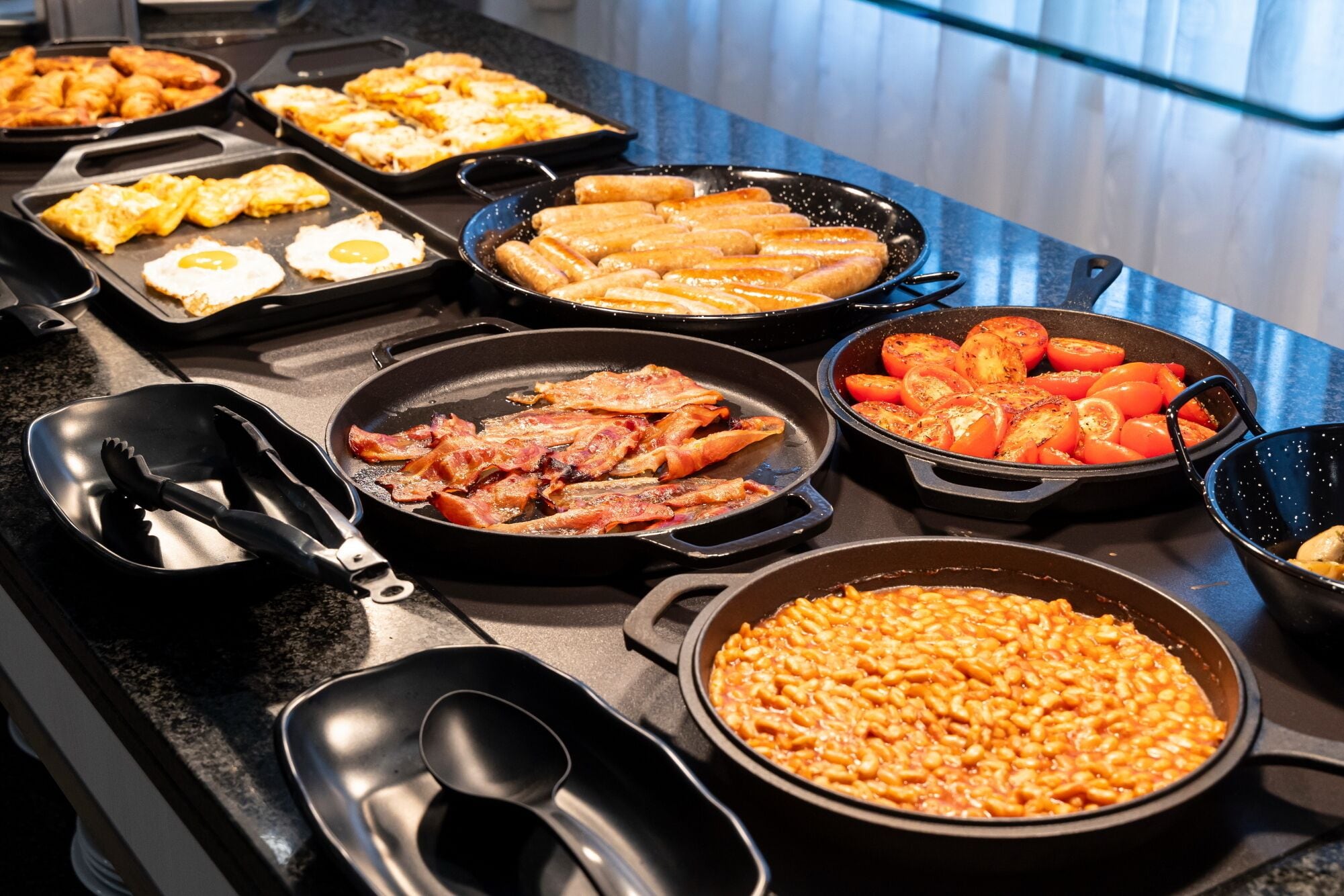 Un bufé con diversos platos, como huevos, tocino, salchichas, tomates y papas al horno.