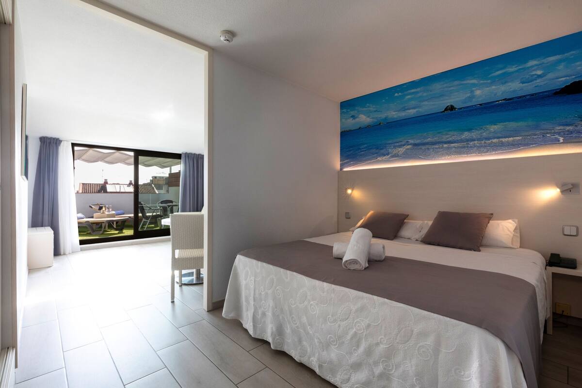 A modern hotel room with a large bed, a painting of a beach scene, and a balcony with outdoor furniture.