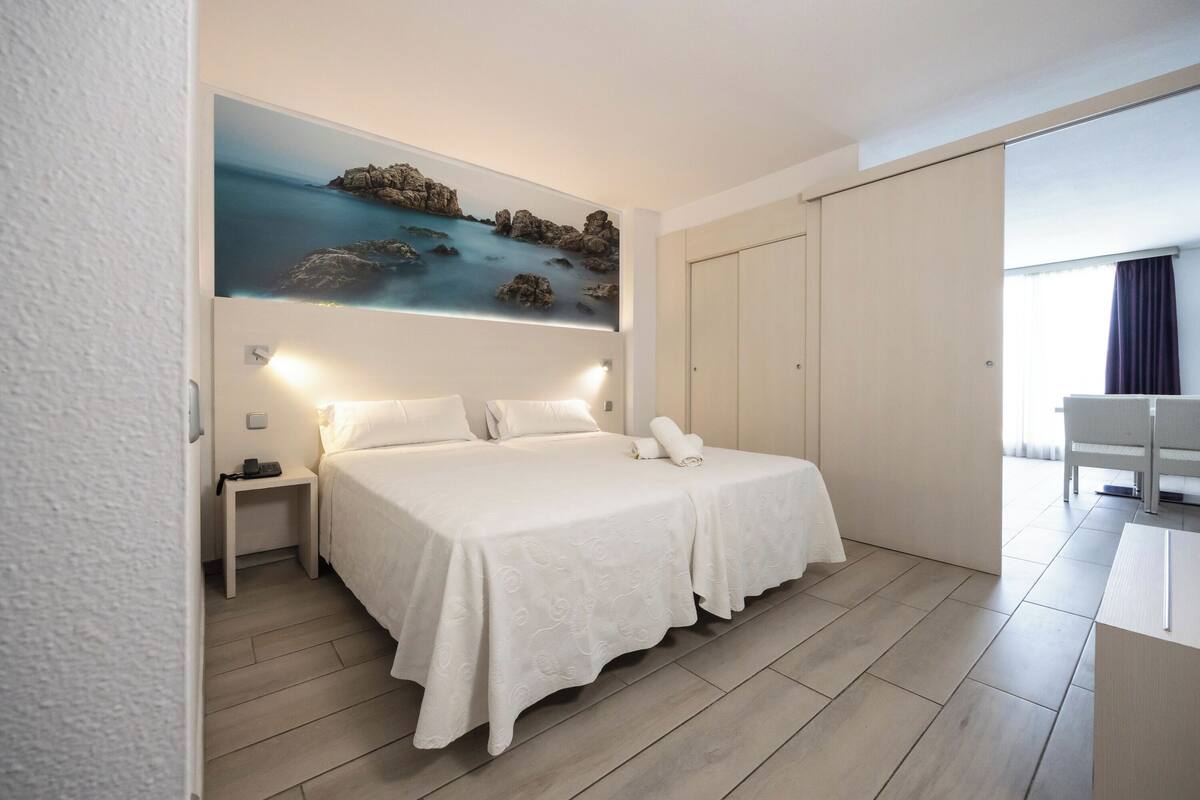 A hotel room with a bed, bedside tables, a painting of a coastal scene, a wardrobe, and a desk with a chair.