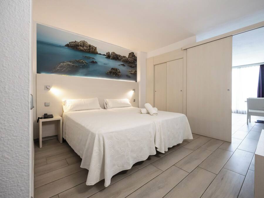 Habitación de hotel con cama, mesitas de noche, un cuadro de una escena costera, armario y escritorio con silla.