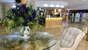 A reception area with a glass-top table, white wicker chair, and a large floral arrangement. - Aparthotel Miami Park (Calella)