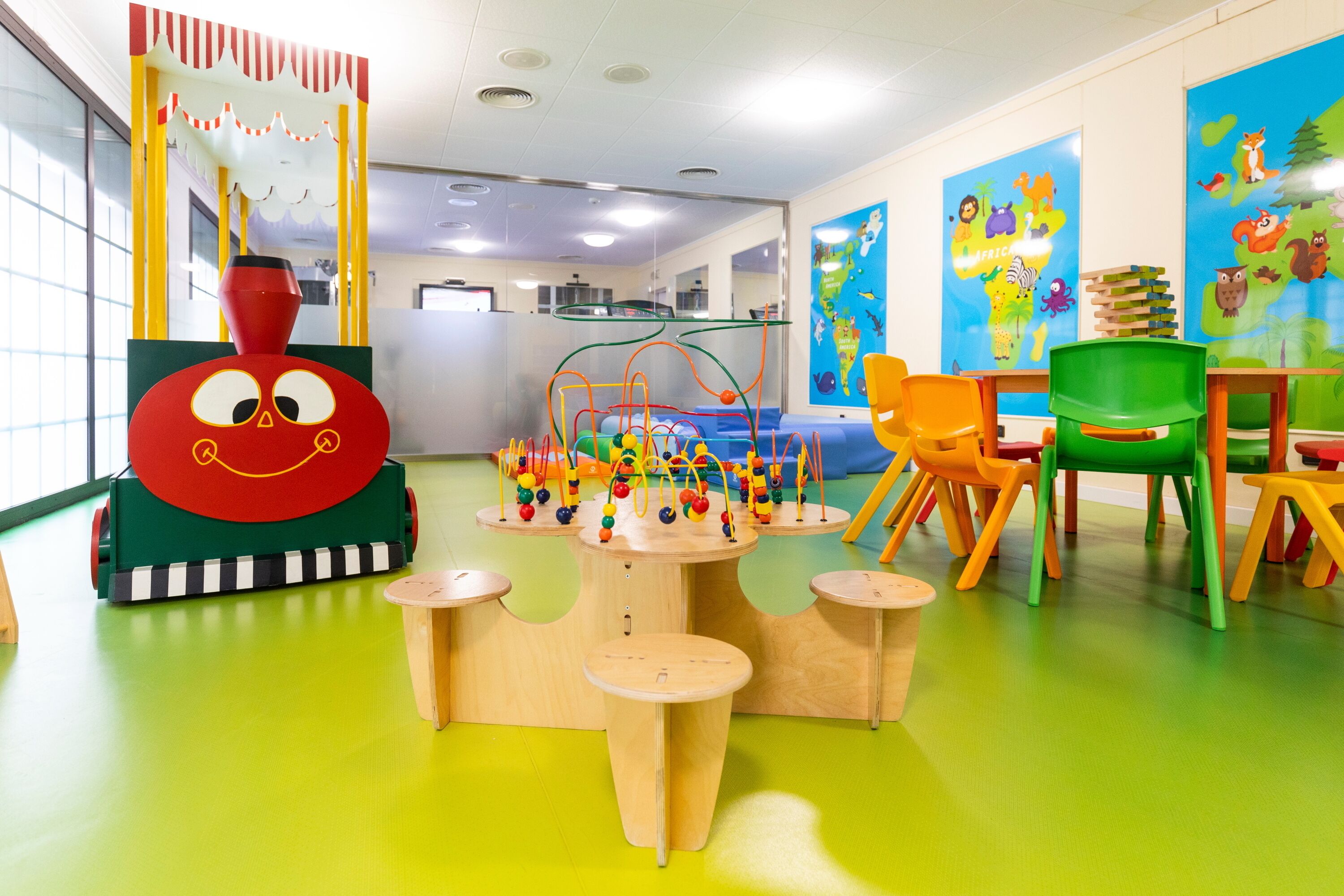A brightly colored children's play area with a large red and green toy train, a play table with colorful toys, and a seating area with orange and green chairs.