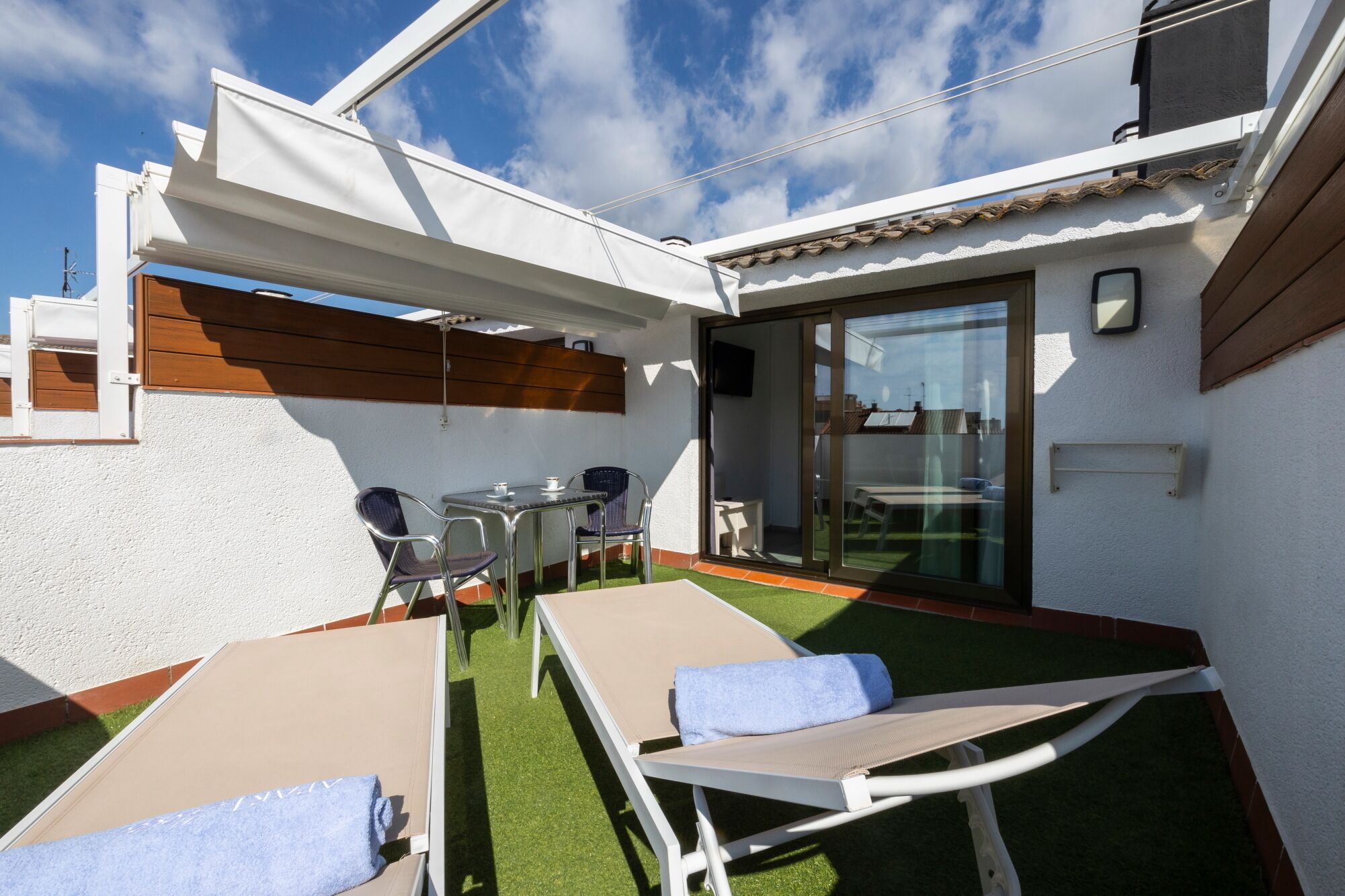 A sunlit outdoor seating area with two lounge chairs and a table, surrounded by white walls and a glass door leading to a covered patio.