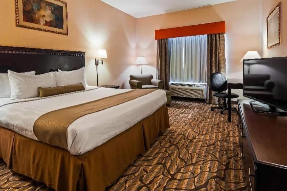 Habitación de hotel con una cama grande, un escritorio con televisor, una silla, una lámpara y un ventanal con cortinas.